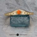 The "Zinnia" band in 14k yellow gold with orange sapphire rests on a rough blue stone against a patterned gray background. Text at the bottom reads "Earth's Treasury.