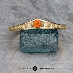 The "Zinnia" band in 14k yellow gold with orange sapphire rests on a rough blue stone against a patterned gray background. Text at the bottom reads "Earth's Treasury.