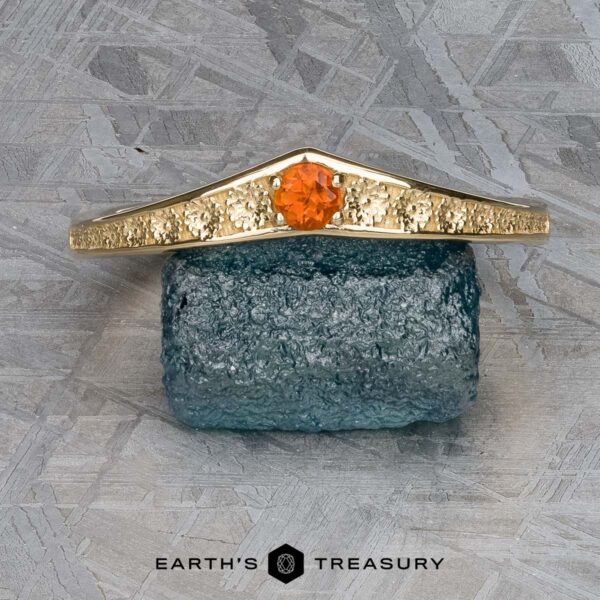The "Zinnia" band in 14k yellow gold with orange sapphire The "Zinnia" band in 14k yellow gold with orange sapphire rests on a rough blue stone against a patterned gray background. Text at the bottom reads "Earth's Treasury.
