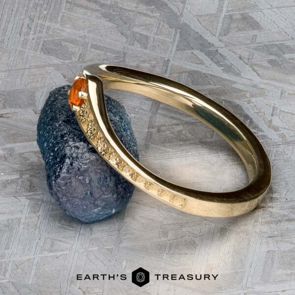 The "Zinnia" band in 14k yellow gold with orange sapphire The "Zinnia" band in 14k yellow gold with orange sapphire sits atop a rough dark mineral on a textured gray surface, featuring Earth's Treasury branding at the bottom.