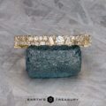 The "Mosaic" band in 14k yellow gold, featuring multiple diamonds, rests on a rough blue gemstone atop a textured gray surface.