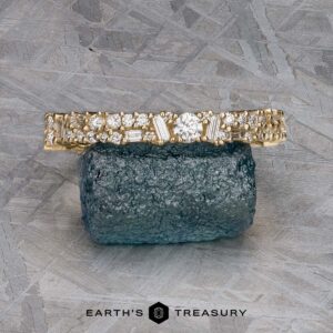 The "Mosaic" band in 14k yellow gold, featuring multiple diamonds, rests on a rough blue gemstone atop a textured gray surface.