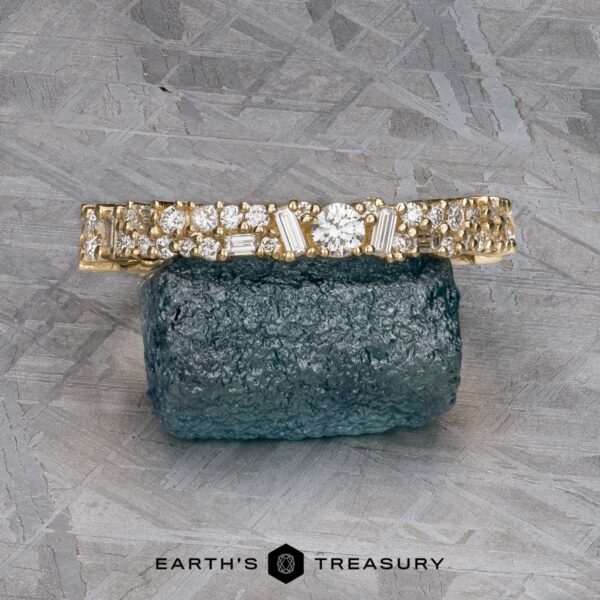 The "Mosaic" band in 14k yellow gold The "Mosaic" band in 14k yellow gold, featuring multiple diamonds, rests on a rough blue gemstone atop a textured gray surface.