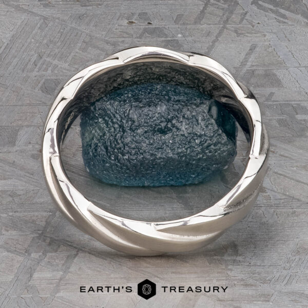 The "Conall" ring in platinum A platinum "Conall" ring with a twisted band encircles a rough blue gemstone, shown on a textured gray surface with "EARTH'S TREASURY" at the bottom.