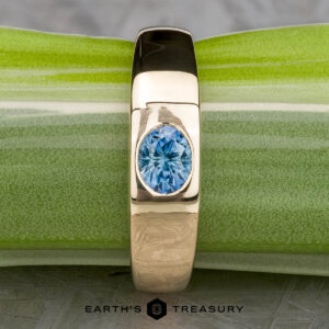 The "Milan" ring in 14k yellow gold with a 1.03-carat Montana sapphire rests on a green and white surface, with The Earth's Treasury logo visible at the bottom.