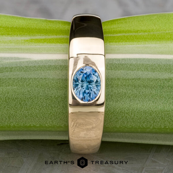 The "Milan" ring in 14k yellow gold with a 1.03-carat Montana sapphire rests on a green and white surface, with The Earth's Treasury logo visible at the bottom.