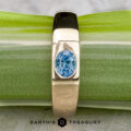 The "Milan" ring in 14k yellow gold with a 1.03-carat Montana sapphire rests on a green and white surface, with The Earth's Treasury logo visible at the bottom.