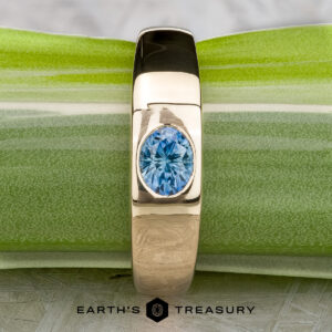 The "Milan" ring in 14k yellow gold with a 1.03-carat Montana sapphire rests on a green and white surface, with The Earth's Treasury logo visible at the bottom.