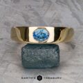 The "Milan" ring in 14k yellow gold with a 1.03-carat Montana sapphire is displayed upright on a raw greenish stone, set against a geometric patterned background with "EARTH'S TREASURY" below.