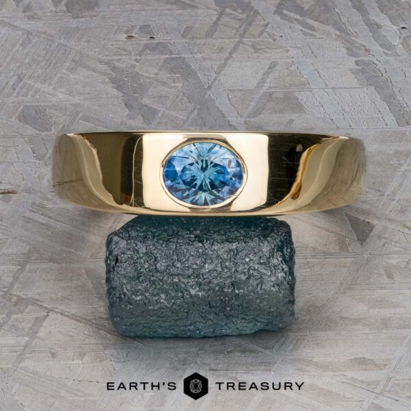 The "Milan" ring in 14k yellow gold with 1.03-carat Montana sapphire The "Milan" ring in 14k yellow gold with a 1.03-carat Montana sapphire is displayed upright on a raw greenish stone, set against a geometric patterned background with "EARTH'S TREASURY" below.