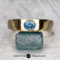 The "Milan" ring in 14k yellow gold with a 1.03-carat Montana sapphire rests on a green and white surface, with The Earth's Treasury logo visible at the bottom.