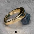 The "Milan" ring in 14k yellow gold with a 1.03-carat Montana sapphire is displayed against a rough blue stone on a textured grey surface, with the "EARTH'S TREASURY" logo and text at the bottom.