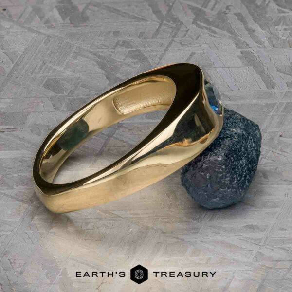 The "Milan" ring in 14k yellow gold with 1.03-carat Montana sapphire The "Milan" ring in 14k yellow gold with a 1.03-carat Montana sapphire is displayed against a rough blue stone on a textured grey surface, with the "EARTH'S TREASURY" logo and text at the bottom.