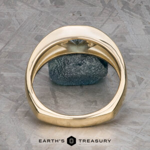 The "Milan" ring in 14k yellow gold with a 1.03-carat Montana sapphire stands upright before a gray stone on a textured surface, with the "Earth's Treasury" logo displayed at the bottom.