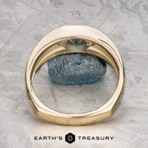 The "Milan" ring in 14k yellow gold with a 1.03-carat Montana sapphire rests on a green and white surface, with The Earth's Treasury logo visible at the bottom.