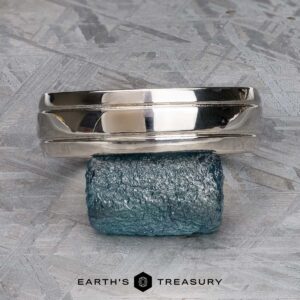 A polished "Helios" Ring in platinum is balanced on a rough blue gemstone, with the "EARTH'S TREASURY" logo and text below on a textured gray surface.