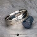 A polished platinum Helios ring is displayed next to a rough blue gemstone on a textured surface, with the text "EARTH'S TREASURY" at the bottom.