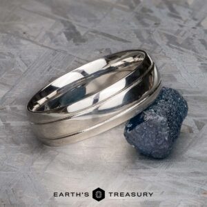 A polished platinum Helios ring is displayed next to a rough blue gemstone on a textured surface, with the text "EARTH'S TREASURY" at the bottom.