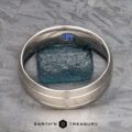 A platinum "Helios" Ring with a small blue gemstone stands upright, encircling a rough blue mineral on a textured grey surface, with the text "EARTH'S TREASURY" displayed below.