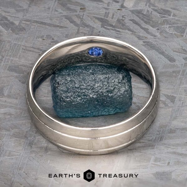 The "Helios" Ring in platinum A platinum "Helios" Ring with a small blue gemstone stands upright, encircling a rough blue mineral on a textured grey surface, with the text "EARTH'S TREASURY" displayed below.