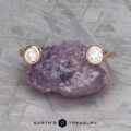 The "Jetty" band in 14k rose gold, featuring two round bezel-set diamonds, rests atop a rough purple gemstone on a textured gray surface. "EARTH'S TREASURY" is displayed at the bottom.