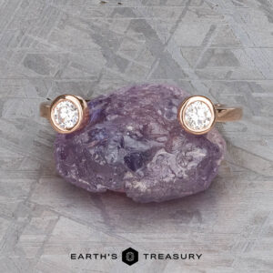 The "Jetty" band in 14k rose gold, featuring two round bezel-set diamonds, rests atop a rough purple gemstone on a textured gray surface. "EARTH'S TREASURY" is displayed at the bottom.