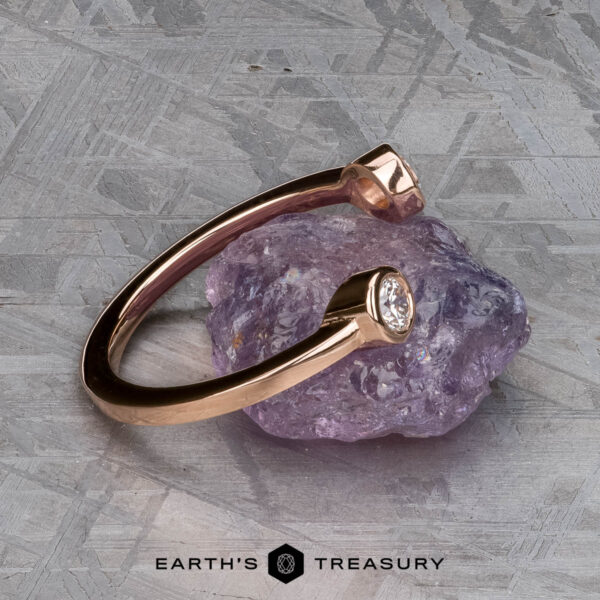 The "Jetty" band in 14k rose gold The "Jetty" band in 14k rose gold, featuring a bezel-set diamond, rests on a rough purple gemstone atop a textured gray surface. The Earth's Treasury logo below highlights the elegance of this unique Jetty-inspired ring design.