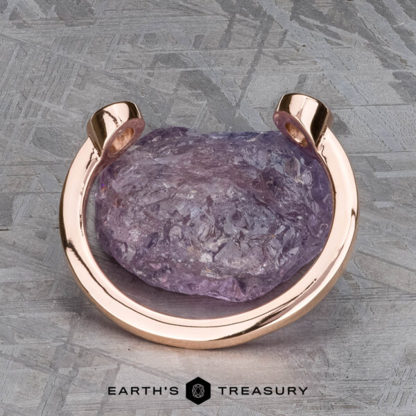 The "Jetty" band in 14k rose gold A rough purple gemstone set in the Jetty band in 14k rose gold rests on a textured gray surface, with the "Earth's Treasury" logo below.