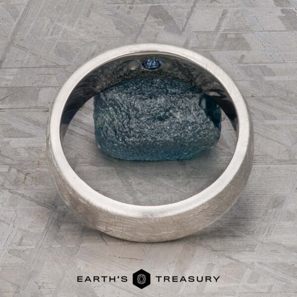 The "Madeira" ring in platinum The "Madeira" ring in platinum with a small blue gemstone is displayed upright on a textured gray surface, the Earth's Treasury logo positioned below.