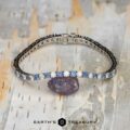 The "Adeline" Yogo Sapphire and Diamond Tennis Bracelet in 14k white gold is shown on a pale, textured surface next to a raw purple stone, with the Earthās Treasury logo featured at the bottom.