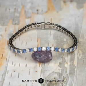 The "Adeline" Yogo Sapphire and Diamond Tennis Bracelet in 14k white gold is shown on a pale, textured surface next to a raw purple stone, with the Earth’s Treasury logo featured at the bottom.