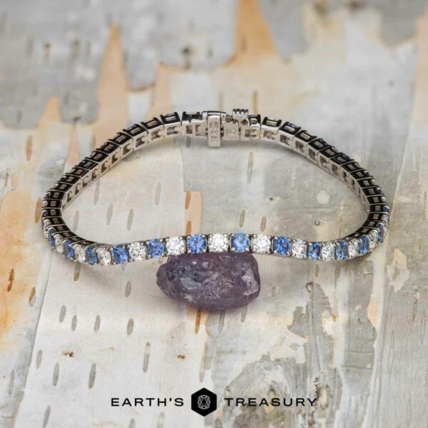 The "Adeline" Yogo Sapphire and Diamond Tennis Bracelet in 14k white gold The "Adeline" Yogo Sapphire and Diamond Tennis Bracelet in 14k white gold is shown on a pale, textured surface next to a raw purple stone, with the Earthās Treasury logo featured at the bottom.