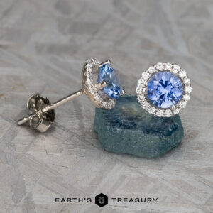 Displayed on a textured gray surface is a pair of stunning earrings known as "Stacked from 15 images. Method=B (R=1,S=3)," featuring heated blue sapphires, each encircled by a halo of small clear stones.