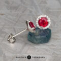 A pair of heated silver stud earrings features vibrant red gemstones as the focal point of the stunning "Stacked from 15 images. Method=B (R=1,S=3)" Ruby Halo Earrings, surrounded by small clear stones and elegantly displayed on a textured surface.