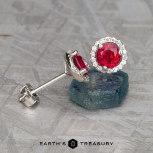 A pair of heated silver stud earrings features vibrant red gemstones as the focal point of the stunning "Stacked from 15 images. Method=B (R=1,S=3)" Ruby Halo Earrings, surrounded by small clear stones and elegantly displayed on a textured surface.