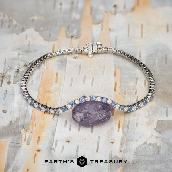 The "Analise" Yogo Sapphire and Diamond Tennis Bracelet in 14k w The "Analise" Yogo Sapphire and Diamond Tennis Bracelet in 14k white gold is shown on a textured, multicolored background. "Earth's Treasury" appears at the bottom.