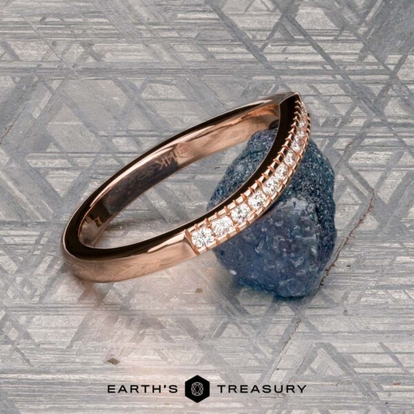 The "Ramona" band in 14K rose gold The "Ramona" band in 14K rose gold rests on a rough blue stone atop a geometric silver background, with the “Earth's Treasury” logo displayed below.
