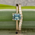 The "Katya" ring in 14K yellow gold with a 0.88-carat Montana sapphire sits with the Contour "Lynn" band in 14K yellow gold on a green leaf. The "Earth's Treasury" logo appears below.