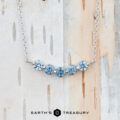 A "Breeze" Necklace in 14K white gold with five round blue gemstones is displayed on a light wood background. The text "EARTH'S TREASURY" appears at the bottom.