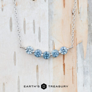 A "Breeze" Necklace in 14K white gold with five round blue gemstones is displayed on a light wood background. The text "EARTH'S TREASURY" appears at the bottom.
