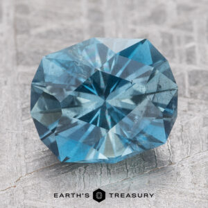 A round, faceted teal Aqua gemstone is placed on a textured surface, with the text "Earth's Treasury" below it.