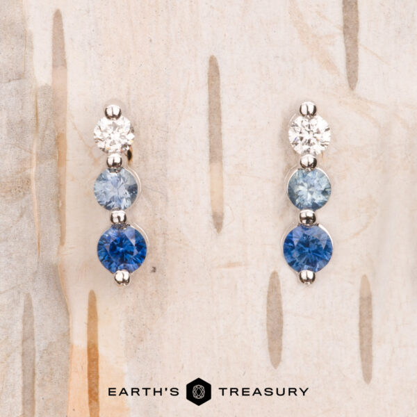 Two pairs of "Dawn" Earrings in 14k white gold, embellished with shimmering white and blue gemstones, sit elegantly on a light wood background. The "Earth's Treasury" logo graces the bottom, accentuating their unique beauty.