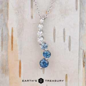 The "Journey" Necklace in 14k white gold features three round blue gemstones and three round white gemstones set in a vertical line, shown on a light wood background.