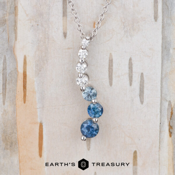 The "Journey" Necklace in 14k white gold The "Journey" Necklace in 14k white gold features three round blue gemstones and three round white gemstones set in a vertical line, shown on a light wood background.