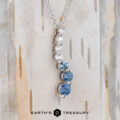 The "Journey" Necklace in 14k white gold features five vertically-set gemstones—three clear and two blue—on a silver chain, displayed on light wood with the Earth’s Treasury logo beneath.