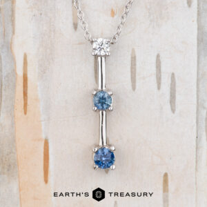 A "Dusk" Necklace pendant in 14k white gold features three vertically aligned gemstones—one clear and two blue—set against a light, textured background. The Earth's Treasury logo appears at the bottom.