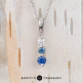 The "Dawn" Necklace in 14k white gold features a vertical pendant with three round stones—white, light blue, and dark blue—shown against a wood background.