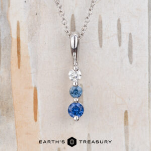 The "Dawn" Necklace in 14k white gold features a vertical pendant with three round stones—white, light blue, and dark blue—shown against a wood background.