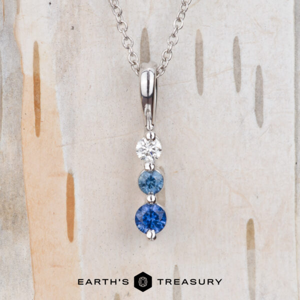 The "Dawn" Necklace in 14k white gold The "Dawn" Necklace in 14k white gold features a vertical pendant with three round stones—white, light blue, and dark blue—shown against a wood background.