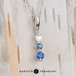 The "Dawn" Necklace in 14k white gold features a vertical pendant with three round gemstones—one white and two blue—displayed against a light textured background.